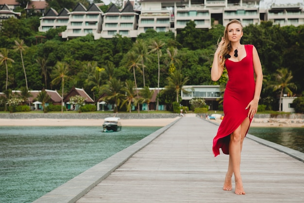 Młoda piękna atrakcyjna kobieta stojąca samotnie na molo w luksusowym hotelu, wakacje, czerwona długa sukienka, blond włosy, seksowna odzież, tropikalna plaża, uwodzicielska, zmysłowa, uśmiechnięta