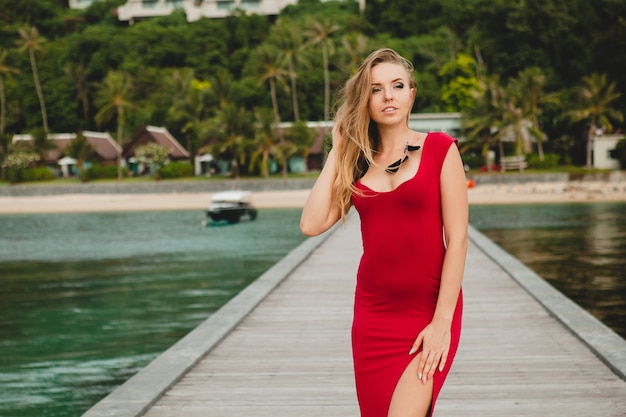 Młoda piękna atrakcyjna kobieta stojąca samotnie na molo w luksusowym hotelu, wakacje, czerwona długa sukienka, blond włosy, seksowna odzież, tropikalna plaża, uwodzicielska, zmysłowa, uśmiechnięta