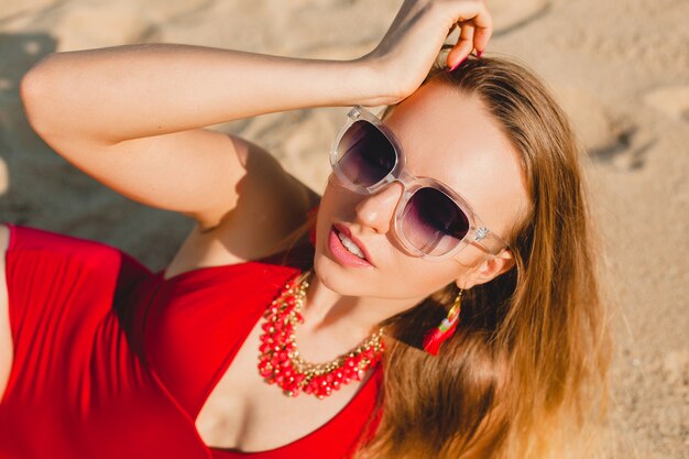Młoda piękna blond kobieta opalając się na piaszczystej plaży w czerwonym kostiumie kąpielowym, okulary przeciwsłoneczne