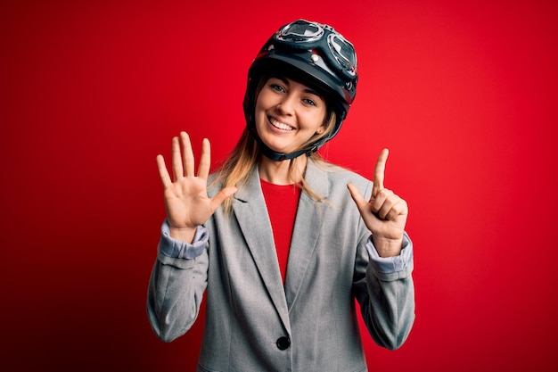 Młoda piękna blond motocyklistka w kasku motocyklowym na czerwonym tle, pokazująca i wskazująca palcami numer siedem, uśmiechając się pewna siebie i szczęśliwa