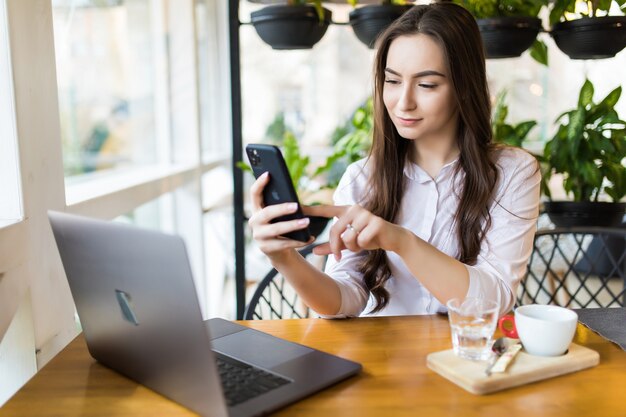 Młoda piękna blondynka trzyma telefon komórkowy siedząc z przenośną netbookiem we wnętrzu kawiarni, urocza marzycielska kobieta korzystająca z telefonu komórkowego i laptopa podczas odpoczynku w kawiarni