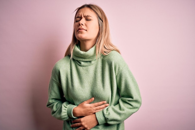 Młoda piękna blondynka ubrana w zimowy wełniany sweter na różowym, odizolowanym tle z ręką na brzuchu, ponieważ nudności bolesna choroba źle się czuje Koncepcja bólu