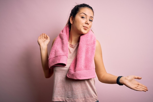 Bezpłatne zdjęcie młoda piękna brunetka sportsmenka ubrana w odzież sportową i ręcznik na różowym tle, nieświadoma i zdezorientowana mina z podniesionymi rękami i rękami koncepcja wątpliwości