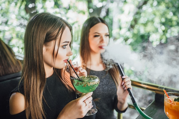 Bezpłatne zdjęcie młoda piękna dziewczyna je zielony koktajl na letnim tarasie nowoczesnej kawiarni, jej dziewczyna pali fajkę wodną.
