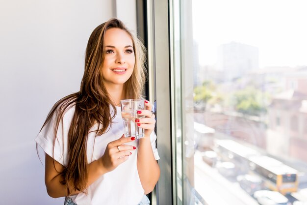 Młoda piękna dziewczyna lub kobieta ze szklanką wody w pobliżu okna w białej koszuli i szarej szacie