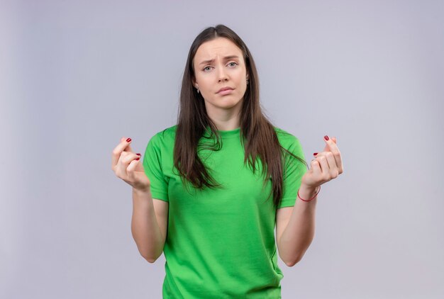 Młoda piękna dziewczyna ubrana w zieloną koszulkę patrząc na kamery niezadowolona, pocierając palce robiąc gest gotówki, prosząc o pieniądze stojąc na białym tle