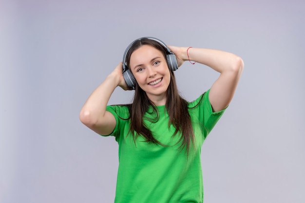Młoda piękna dziewczyna ubrana w zielony t-shirt ze słuchawkami uśmiechnięta pozytywna i szczęśliwa ciesząc się ulubioną muzyką stojącą na białym tle