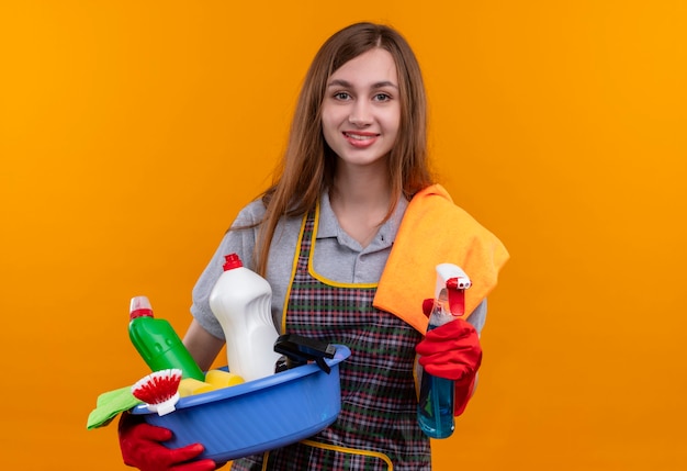 Młoda piękna dziewczyna w fartuch i rękawice gumowe, trzymając umywalkę z narzędziami do czyszczenia i spray z dywanikiem uśmiechnięty szczęśliwy i pozytywny