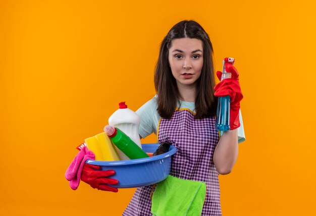 Młoda piękna dziewczyna w fartuch i rękawice gumowe, trzymając umywalkę z narzędziami do czyszczenia i sprayem do czyszczenia, wygląda pewnie, gotowa do czyszczenia