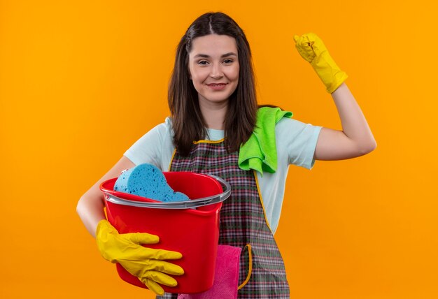 Młoda piękna dziewczyna w fartuch i rękawice gumowe, trzymając wiadro z narzędziami do czyszczenia, podnosząc pięść jak uśmiechnięty zwycięzca