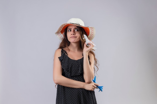 Młoda piękna dziewczyna w sukience w groszki w letnim kapeluszu trzyma samoloty-zabawki, patrząc na bok niezadowolony z sceptycznym wyrazem twarzy stojącej na białym tle