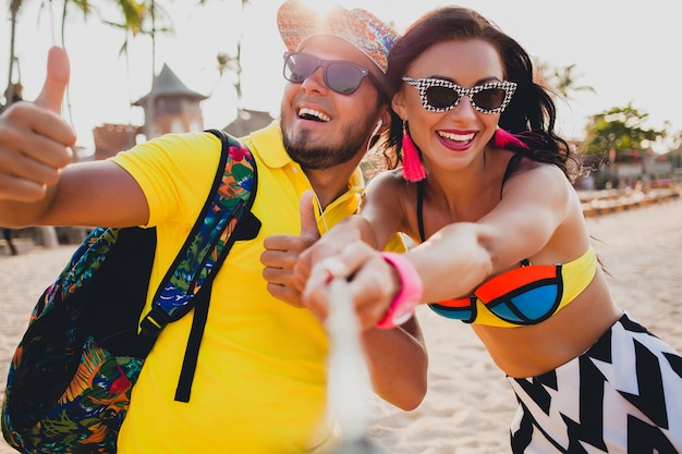 Młoda piękna hipster para zakochana na tropikalnej plaży, robienie zdjęć selfie na smartfonie, okulary przeciwsłoneczne, stylowy strój, letnie wakacje, zabawa, uśmiechnięty, szczęśliwy, kolorowy, pozytywne emocje