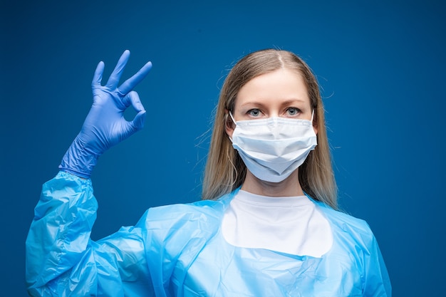 Młoda piękna kaukaska kobieta w niebieskiej sukni medycznej z białą maską medyczną na twarzy patrzy w kamerę i pokazuje OK