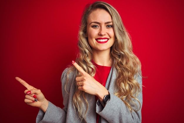 Bezpłatne zdjęcie młoda piękna kobieta biznesu ubrana w elegancką kurtkę, stojąca na czerwonym, odizolowanym tle, uśmiechnięta i patrząca na kamerę wskazującą dwiema rękami i palcami na bok