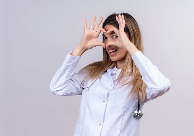 Młoda piękna kobieta lekarz ubrana w biały fartuch ze stetoskopem robi romantyczny gest serca palcami patrząc przez ten znak jest piękny