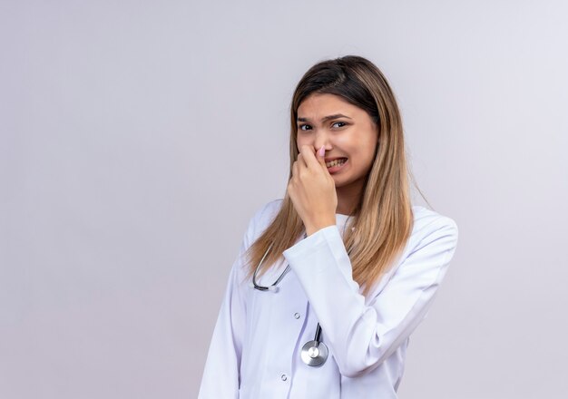 Młoda piękna kobieta lekarz ubrana w biały fartuch ze stetoskopem, zamykając nos palcami czując przyjemny zapach