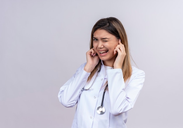 Młoda piękna kobieta lekarz ubrana w biały fartuch ze stetoskopem zamykając uszy palcami z zirytowanym wyrazem hałasu głośnego dźwięku