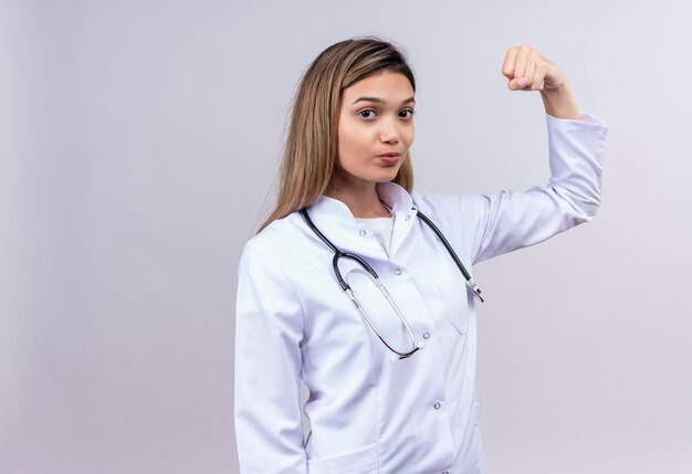 Młoda piękna kobieta lekarz ubrany w biały fartuch ze stetoskopem, patrząc pewnie podniesionej pięści, udając zwycięzcę