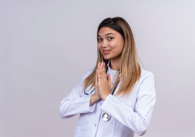 Młoda piękna kobieta lekarz ubrany w biały fartuch ze stetoskopem, trzymając rękę w geście namaste modlitwy, wdzięczny i szczęśliwy