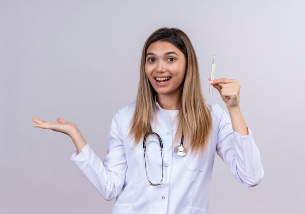 Młoda piękna kobieta lekarz ubrany w biały fartuch ze stetoskopem trzymając termometr patrząc pewnie, prezentując z ramieniem oh jej dłoni