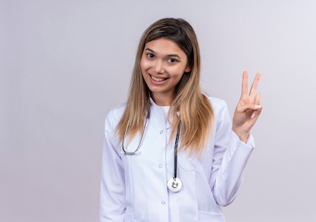 Młoda piękna kobieta lekarz ubrany w biały fartuch ze stetoskopem uśmiechnięty przyjazny pokazując znak zwycięstwa