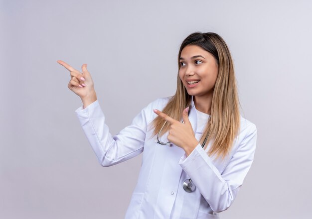 Młoda piękna kobieta lekarz ubrany w biały fartuch ze stetoskopem, wskazując palcami wskazującymi na bok uśmiechnięty przyjazny