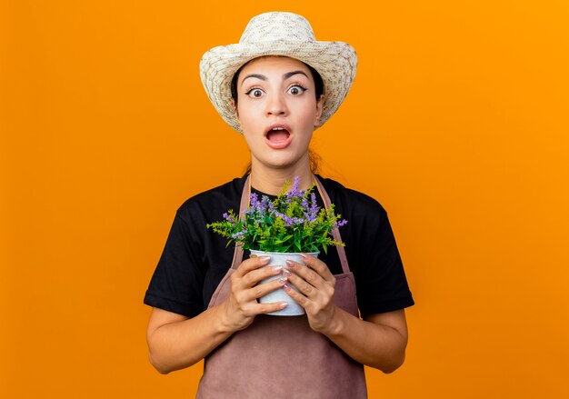 Młoda piękna kobieta ogrodnik w fartuch i kapelusz gospodarstwa roślina doniczkowa patrząc na przód zaskoczony stojąc nad pomarańczową ścianą