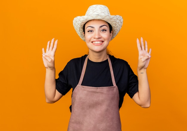 Młoda piękna kobieta ogrodnik w fartuch i kapelusz pokazując i wskaż palcami numer osiem stojąc na pomarańczowej ścianie