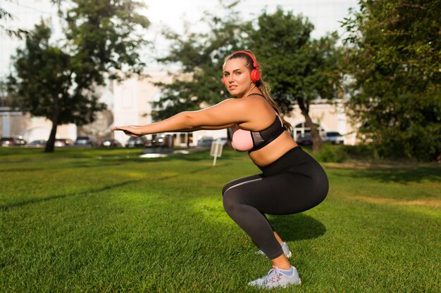 Młoda piękna kobieta plus size w sportowym topie i legginsach z czerwonymi słuchawkami marzycielsko patrząc w kamerę podczas ćwiczeń sportowych na trawie w parku miejskim