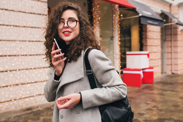 Młoda piękna kobieta rozmawia na smartfonie, jesienny styl miasta, ciepły płaszcz, okulary, szczęśliwa, uśmiechnięta, trzymając telefon w dłoni, kręcone włosy