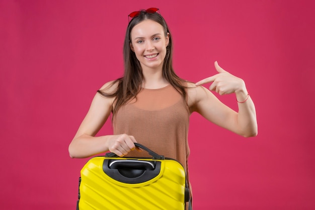 Młoda piękna kobieta stojąca z walizką podróżną, wskazując palcem na siebie, uśmiechając się z radosną buźką na różowym tle
