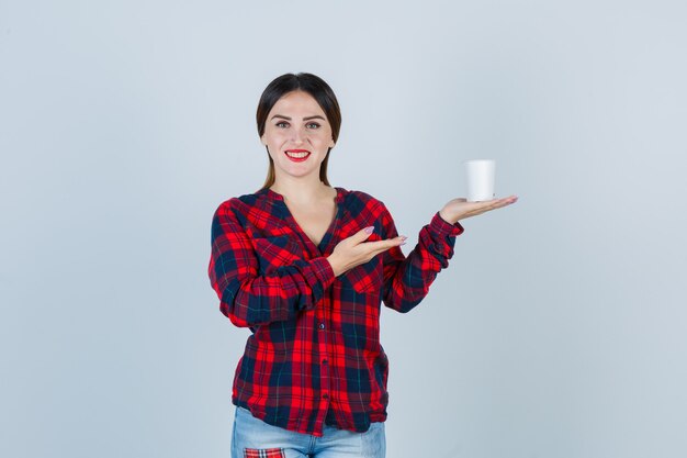 Młoda piękna kobieta trzyma plastikową szklankę, pokazując powitalny gest w casualowej koszuli, dżinsach i patrząc radosny, widok z przodu.