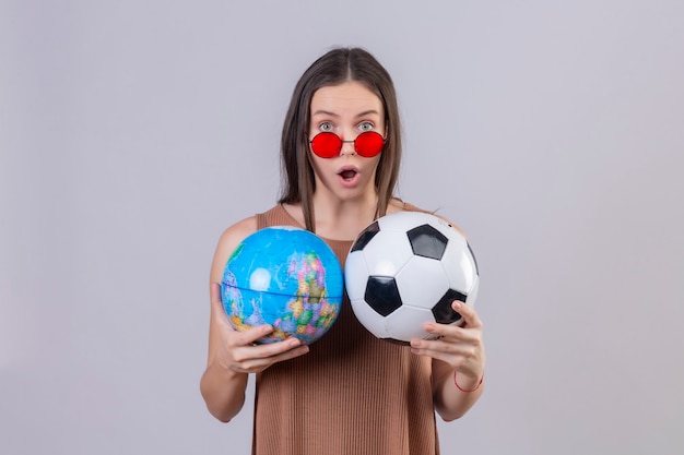 Młoda piękna kobieta ubrana w czerwone okulary przeciwsłoneczne, trzymając piłkę nożną i kula ziemska zdumiony i zaskoczony, stojąc na białym tle