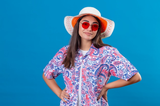Młoda piękna kobieta ubrana w letni kapelusz i czerwone okulary przeciwsłoneczne z pewnym uśmiechem na niebieskiej ścianie