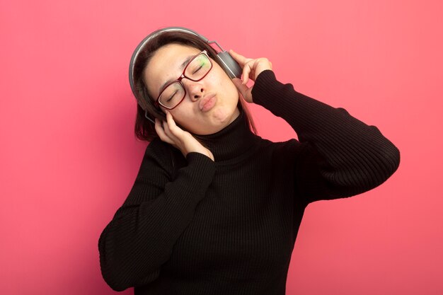 Młoda piękna kobieta w czarnym golfie i okularach ze słuchawkami, ciesząc się ulubioną muzyką z zamkniętymi oczami stojąc na różowej ścianie