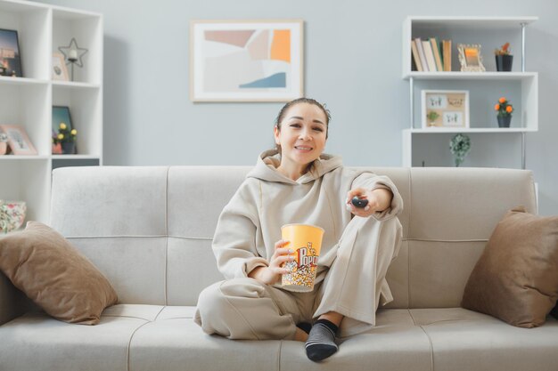 Młoda piękna kobieta w domowych ubraniach, siedząc na kanapie w domu, z wiadrem popcornu, trzymając pilota zdalnego oglądania telewizji, będąc szczęśliwym i pozytywnym uśmiechniętym przyjaznym