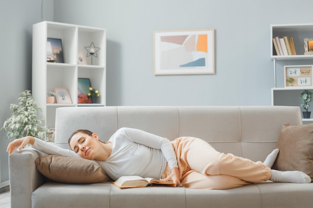 młoda piękna kobieta w domowych ubraniach śpi na kanapie w domu wnętrze z książką spędzając weekend w domu