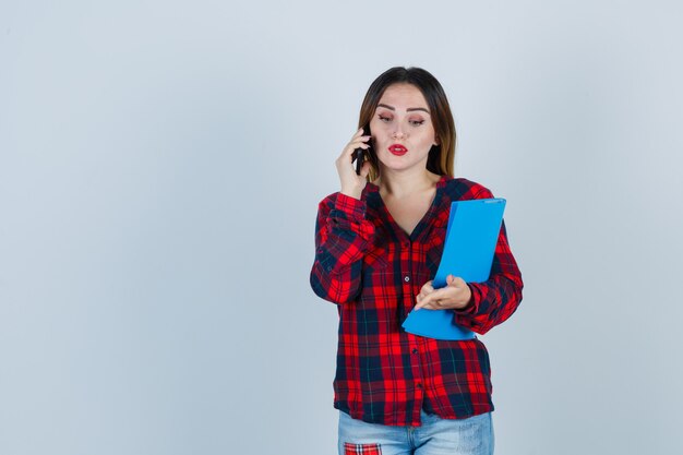 Młoda piękna kobieta w dorywczo koszula, dżinsy trzymając folder podczas rozmowy przez telefon, wskazując w dół i patrząc niezdecydowany, widok z przodu.