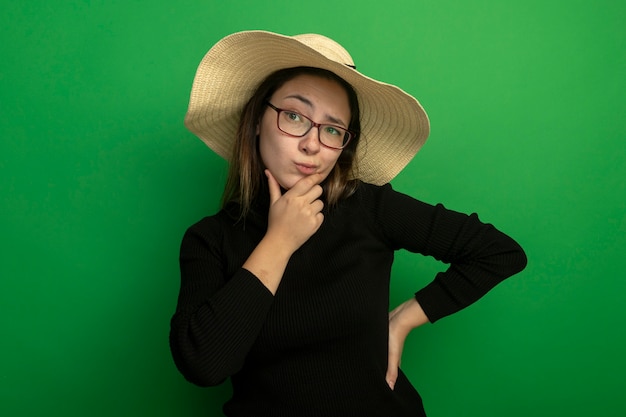 Młoda piękna kobieta w letnim kapeluszu w czarnym golfie i okularach patrząc na bok ze sceptycznym wyrazem twarzy z ręką na brodzie, myśląc stojąc nad zieloną ścianą