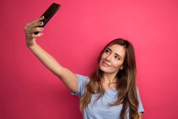 Młoda piękna kobieta w niebieskiej koszulce za pomocą smartfona robi selfie uśmiechając się radośnie stojąc nad różowym