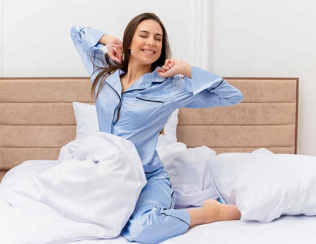 Młoda piękna kobieta w niebieskiej piżamie siedzi na łóżku, budząc się, rozciągając ręce, ciesząc się porannym czasem we wnętrzu sypialni na jasnym tle