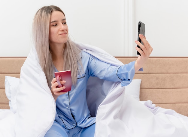 Młoda piękna kobieta w niebieskiej piżamie siedzi na łóżku pod blenketem z filiżanką kawy robi selfie za pomocą smartfona szczęśliwa i pozytywna we wnętrzu sypialni na jasnym tle