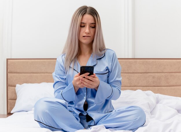 Młoda piękna kobieta w niebieskiej piżamie siedzi na łóżku przy użyciu smartfona, patrząc zdezorientowany gryzienie wargi we wnętrzu sypialni na jasnym tle