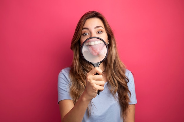 Młoda piękna kobieta w niebieskim t-shirt trzyma szkło powiększające przed jej ustami wystający język zabawy stojąc na różowym tle