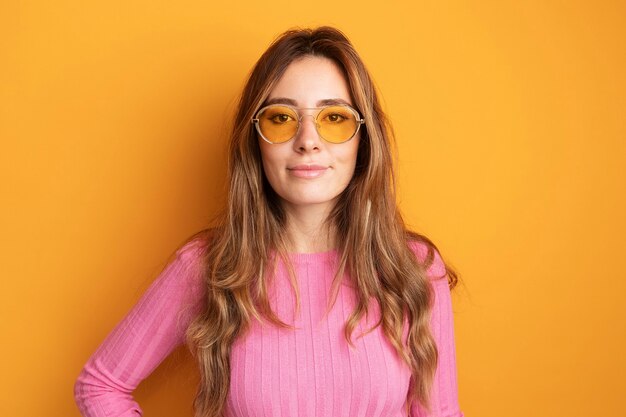 Młoda piękna kobieta w różowym topie w okularach, patrząc na kamerę, uśmiechając się pewnie, stojąc nad pomarańczą