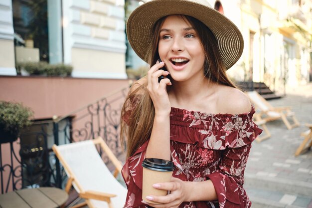 Młoda piękna kobieta w sukience i słomkowym kapeluszu trzymająca w ręku filiżankę kawy do wyjścia i rozmawiającą na telefonie komórkowym, jednocześnie szczęśliwie patrząc na bok na przytulnej ulicy miasta