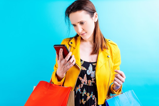 Młoda piękna kobieta w żółtej kurtce na niebieskim tle zamyślony patrząc na smartphone z torby na zakupy. koncepcja zakupoholiczka ofiara mody