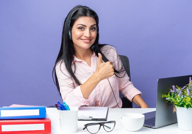Młoda piękna kobieta w zwykłych ubraniach na sobie zestaw słuchawkowy z mikrofonem, patrząc uśmiechnięty pewnie pokazując kciuk do góry, siedząc przy stole z laptopem na niebieskim tle, pracując w biurze