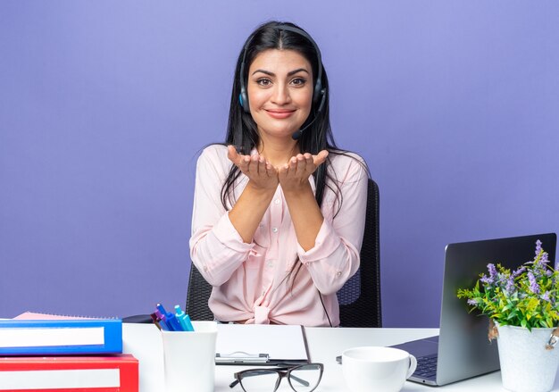Młoda piękna kobieta w zwykłych ubraniach na sobie zestaw słuchawkowy z mikrofonem szczęśliwa i pozytywnie trzymająca się za ręce przed sobą, siedząca przy stole z laptopem nad niebieską ścianą, pracująca w biurze