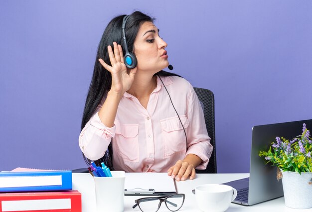 Młoda piękna kobieta w zwykłych ubraniach na sobie zestaw słuchawkowy z mikrofonem, trzymając rękę nad uchem, próbując słuchać, siedząc przy stole z laptopem nad niebieską ścianą, pracując w biurze